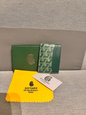 Grön korthållare från G - Snygg grön korthållare från G med klassiskt mönster och guldfärgad logga. Tillverkad i exklusivt läder med plats för flera kort. Kommer med originalask, putsduk och kort. Perfekt accessoar för dig som gillar lyxiga detaljer.