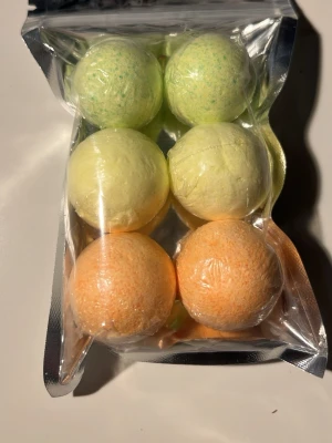 Färgglada badbomber i presentförpackning - Sex badbomber i olika färger – grönt, gult och orange – i en genomskinlig plastförpackning. Badbomberna är runda och kompakta, perfekta för ett lyxigt bad. Passar dig som vill ha en avslappnande stund med doft och färg i badet.