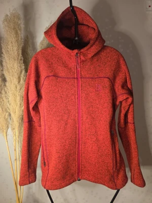 Röd hoodie från Haglöfs med dragkedja - Säljer en röd fleece hoodie från Haglöfs med hel dragkedja och två fickor med dragkedja. Materialet är stickat och känns mjukt och varmt. Hoodien har en huva och diskret Haglöfs-logga på bröstet. Perfekt för kalla dagar.