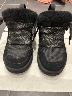 Svarta sneakers från UGG med päls - Säljer ett par svarta sneakers från UGG med fluffig päls runt öppningen och grova svartvita snören. Skorna har en robust sula och är tillverkade i mesh och syntetmaterial. Perfekt för dig som vill ha något unikt och bekvämt.