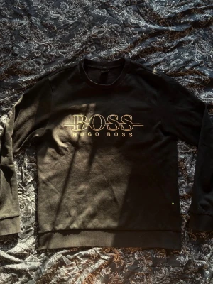 Svart sweatshirt från Hugo Boss - Svart sweatshirt från Hugo Boss med stort guldfärgat BOSS-tryck på bröstet. Klassisk rund halsringning och ribbade muddar vid ärmslut och nederkant. Perfekt för en stilren och avslappnad look.