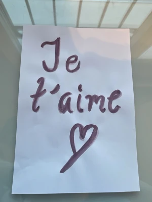 Handskrivet kärleksbrev på papper - Ett vitt papper med handskriven text i lila färg där det står 'Je t'aime' och en hjärtsymbol under. Perfekt som en personlig hälsning eller dekoration. Pappret har en enkel och charmig design.