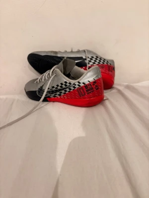 Nike Mercurial silver/röd fotbollsskor - Snygga Nike Mercurial fotbollsskor i silver med svarta och röda detaljer. Skorna har schackrutigt mönster på sidan, svart sula och vita skosnören. Tillverkade i syntetmaterial för bra grepp och känsla på planen. Perfekta för dig som vill sticka ut på fotbollsplanen.