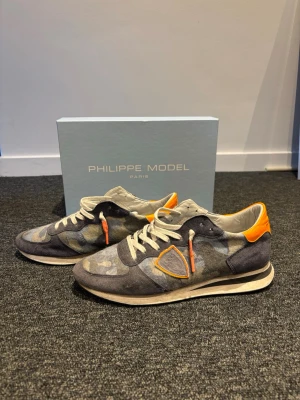 Philippe Model grå-orange sneakers - Snygga sneakers från Philippe Model i grått camouflagemönster med orange detaljer och vita skosnören. Nypris 2799 och mitt pris endast 999. Skick 7/10