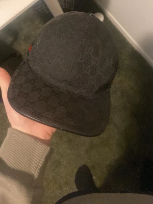 Svart keps från Gucci med monogram - Snygg svart keps från Gucci med klassiskt GG-monogram över hela kepsen. På sidan finns en ikonisk grön och röd rand. Kepsen har böjd skärm och justerbar rem bak. Materialet är slitstark bomullscanvas som ger en exklusiv känsla.