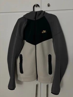 Nike hoodie i grått och beige - Nike hoodie med dragkedja i grått, beige och svart. Snygg kontrast mellan färgerna och gul Nike-logga på bröstet. Två fickor med dragkedja framtill och huva. Perfekt för chill eller träning. Mjuk och skön materialkänsla.