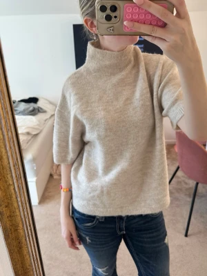 Beige stickad polo tröja - En beige stickad tröja med polo och korta ärmar. Tröjan har en avslappnad passform och ribbstickade detaljer vid halsen och nederkanten. Perfekt för lager-på-lager och enkel att matcha med jeans eller kjol.