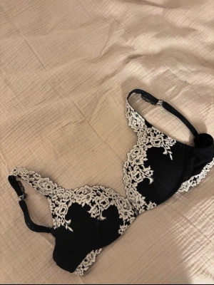 Svart intimissimi bh med vit spets - Snygg svart bh med detaljerad vit spetsbrodyr över kuporna och banden. Bh:n har vadderade kupor och justerbara axelband. Materialet är en kombination av mesh och spets som ger en elegant och feminin look.