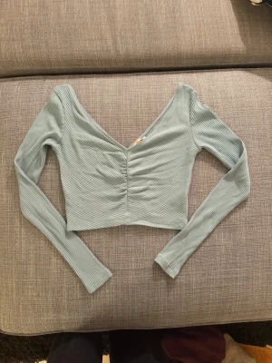  Långärmad  crop top - Trendig  crop top med långa ärmar och djup V-ringning. Toppen har ribbad struktur och snygg rynkning framtill som ger en cool detalj. Perfekt att matcha med  jeans eller kjol för en modern look.