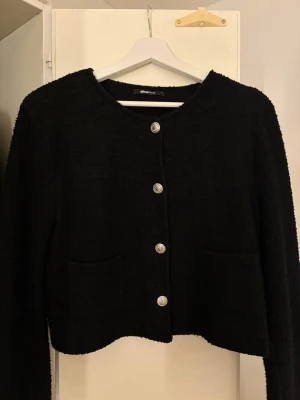 Svart tweedkofta med silverknappar - Stilren svart tweedkofta från Gina Tricot med rund halsringning och dekorativa silverfärgade knappar framtill. Koftan har två praktiska fickor framtill och en croppad passform som ger en trendig look. Perfekt att matcha med jeans eller kjol. Passar som xs 
