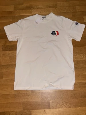 Vit Moncler t-shirt med logotyp - Vit t-shirt från Moncler med klassisk rund hals och korta ärmar. På bröstet finns Monclers ikoniska logotyp i blått och rött, och en liten logotyp på ena ärmen. Materialet är mjukt och bekvämt, perfekt för en stilren look.