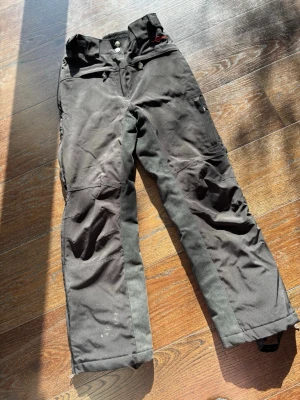 svarta ridtäck byxor  - Säljer ett par svarta täckbyxor från Fjällräven med förstärkta knän och grå detaljer längs benen. Byxorna har flera fickor med dragkedja och knappstängning samt hällor för bälte. Perfekta för kalla dagar och utomhusaktiviteter.