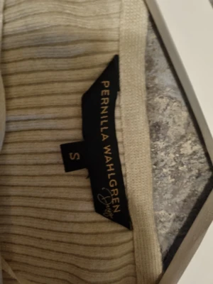 Beige omlottopp Pernilla Wahlgren-oanvänd  - Beige ribbad stickad omlottopp från Pernilla Wahlgren. Långärmad modell med v-ringning och diskret metallogo vid nederkanten. Perfekt för dig som gillar stilrena och bekväma plagg.