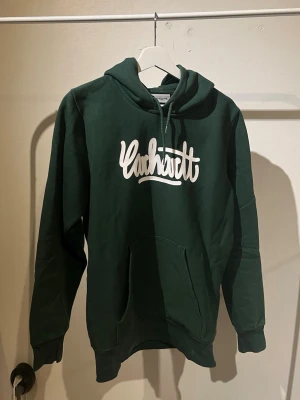 Mörkgrön Carhartt hoodie - Snygg mörkgrön hoodie från Carhartt med stor vit logga på bröstet. Klassisk känguruficka framtill och justerbar huva med dragsko. Tillverkad i mjuk bomullsblandning som känns skön mot huden. Perfekt för en avslappnad streetwear-look.