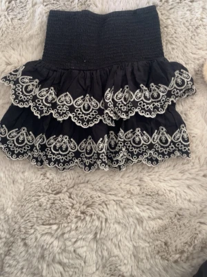 Svart volangkjol med broderi - Supersöt svart kortkjol med två lager volanger och vitt broderat mönster längs kanterna. Kjolen har bred smock i midjan för skön passform och ger en riktigt trendig look.