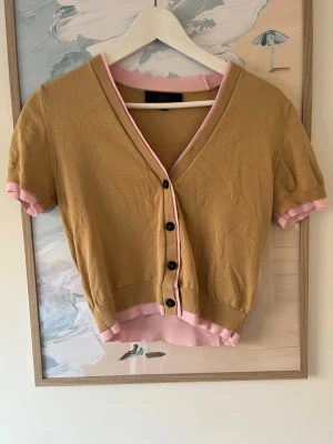 Kortärmad kofta från Zara i brunt och rosa - Snygg kortärmad kofta från Zara i brunt med rosa detaljer vid ärmslut, nederkant och halsringning. Koftan har v-ringning och svarta knappar framtill. Perfekt för lager-på-lager och en trendig look.
