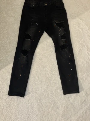 Svarta ripped jeans med färgstänk - Säljer ett par svarta jeans med stora slitningar på båda benen och coola färgstänk i orange och vitt. Jeansen har smal passform och klassisk femficksdesign. Perfekta för dig som gillar en edgy streetstil.