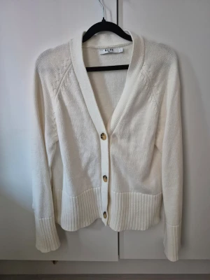 Vit kofta från NA-KD med knappar - Säljer en krämvit kofta från NA-KD i storlek S. Koftan har djup V-ringning, tre stora knappar framtill och breda ribbade muddar vid ärmslut och nederkant. 