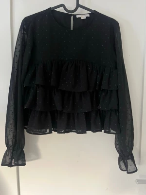 Svart volangblus med prickigt mönster - Svart blus med transparenta långärmar och små prickar i tyget. Blusen har flera lager volanger framtill och bak Den är ifrån Gina tricot och är i storlek 158/164