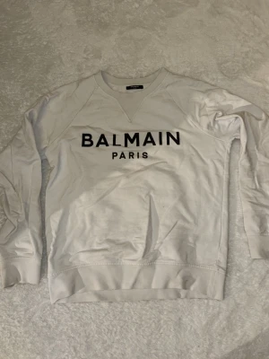Vit sweatshirt från Balmain Paris - Snygg vit sweatshirt från Balmain Paris med svart logotyptryck på bröstet. Tröjan har rund halsringning, långa ärmar och ribbade muddar vid ärmslut och nederkant. Perfekt för en trendig och avslappnad stil.