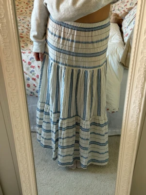 Vit och blå långkjol med ränder - Långkjol i vitt med blåa vertikala och horisontella ränder. Kjolen har en avslappnad passform och är sydd i flera volanger för en bohemisk känsla. Perfekt för varmare dagar och enkel att styla med en enkel topp.