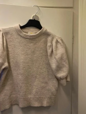 Beige stickad tröja med puffärm - Supersöt beige stickad tröja med trendiga puffärmar och ribbade muddar vid ärmslut och nederkant. Tröjan har rund halsringning och är gjord i ett mjukt material som känns skönt mot huden. Perfekt för dig som gillar en stilren men ändå unik look.