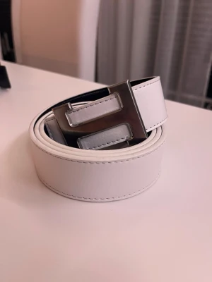 Vitt läderbälte med H-spänne - Snyggt vitt bälte i läder med ett stort, silverfärgat H-format metallspänne. Bältet har stilrena sömmar och en klassisk design som passar till jeans eller byxor. Perfekt accessoar för att lyfta din outfit.