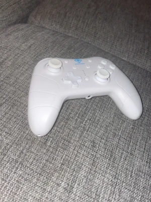 Nintendo Switch Bluetooth Controller - Snygg vit trådlös handkontroll med ergonomisk design och vita joysticks. Passar perfekt för gaming och har tydliga knappar samt styrkors. Tillverkad i plast och har en modern look med blå detalj på ovansidan. Funkar både till Nintendo Switch och Nintendo Switch 2.