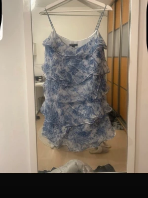 Blåvit volangklänning med smala band - Söt kort klänning med blått och vitt blommönster, tunna axelband och flera lager volanger. Perfekt för sommarens varma dagar. Tillverkad i lätt och luftig polyester för en bekväm känsla.