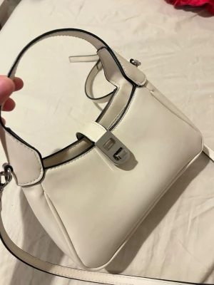 Vit axelremsväska i läderlook - Stilren handväska i krämvit färg med både handtag och justerbar axelrem. Väskan har silverfärgat spänne framtill och svarta kanter som ger en snygg kontrast. Tillverkad i slät konstläder och har en modern, boxig form.