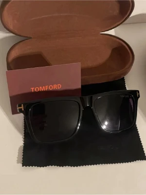 Svarta Tom Ford solglasögon - Snygga solglasögon från Tom Ford med klassisk rektangulär form och diskreta guldfärgade detaljer vid bågarna. Glasögonen kommer med originalfodral och putsduk, perfekt för dig som vill ha en stilren accessoar.