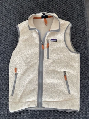 Vit fleeceväst från Patagonia - Säljer en krämvit fleeceväst från Patagonia med grå dragkedjor och detaljer i läder. Västen har hög krage, tre fickor med dragkedja och mjukt teddyfoder. Perfekt för lager-på-lager och riktigt snygg till jeans eller hoodies.