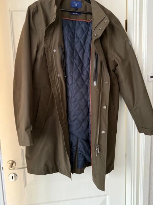 Olivgrön rock från Gant- the city rain coat! - Olivgrön rock från Gant med quiltat blått innerfoder och diskret logotyp på ärmen. Rocken har hög krage, dragkedja och knappar framtill samt flera praktiska fickor. Supersnygg och fräsch, nästan oanvänd!