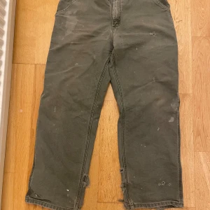 Carhartt byxor/work pants - Säljer ett par gröna cargobyxor från Carhartt med bred passform och flera praktiska fickor både fram och bak. Byxorna har slitna detaljer och råa benslut.  Midjemått halvvägs: 42 cm. Längs utsida: 100 cm