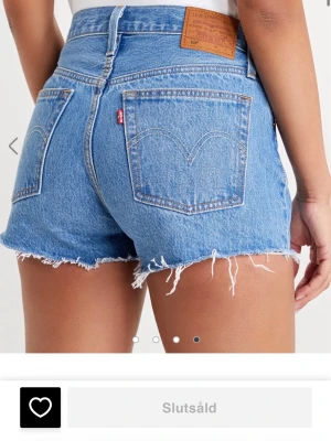 Levi's blå högmidjade jeansshorts - Klassiska blå jeansshorts från Levi's med hög midja och fransiga benslut. Shortsen har fem fickor, knappgylf och den ikoniska Levi's-lappen bak i midjan. Perfekta för en avslappnad och trendig sommarstil. Slutsålda online och säljer då de tyvärr är för små. Använda endast två gånger. Storlek W28