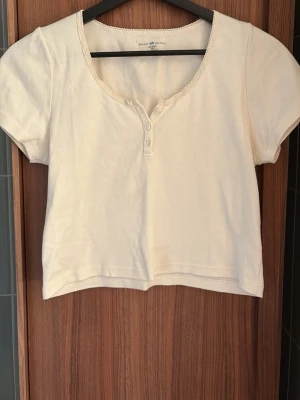 Vit croppad topp från Brandy Melville - Söt vit croppad topp från Brandy Melville med korta ärmar och rundad halsringning. Toppen har dekorativa knappar framtill och fina spetsdetaljer längs halsen. Tillverkad i mjuk bomull, perfekt för en avslappnad och trendig look❤️❤️