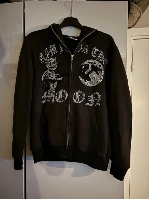 Svart hoodie med strass och tryck - Säljer en svart hoodie med dragkedja och huva. På framsidan finns ett stort tryck med figurer. Hoodien har en avslappnad passform. Perfekt för dig som gillar streetwear och statement-plagg. Nackdelen är att många stenar har ramlat av men det är inget man tänker på så mycket. 