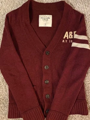 Vinröd kofta från Abercrombie & Fitch - Vinröd kofta med knappar från Abercrombie & Fitch. Har broderad A&F-logga och vita ränder på ena ärmen. Djup V-ringning, ribbade muddar och två fickor framtill. Perfekt för lager-på-lager och snygg till jeans.