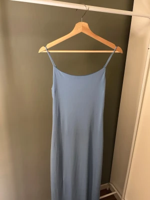 Ljusblå långklänning med smala band - Säljer en enkel och stilren ljusblå långklänning med smala axelband. Klänningen har en rak passform och är tillverkad i ett mjukt tyg som faller snyggt längs kroppen. Perfekt för varma dagar eller sommarens alla tillfällen.