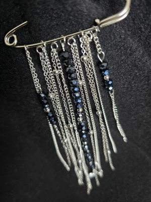 Dekorativ säkerhetsnål med kedjor - Unik säkerhetsnål i silverfärg med hängande kedjor och svarta glaspärlor. Nålen har flera tunna kedjor i olika längder, vissa dekorerade med små metallkulor och en liten trollsländor. Perfekt accessoar till skjorta, tröja, byxor eller jacka.