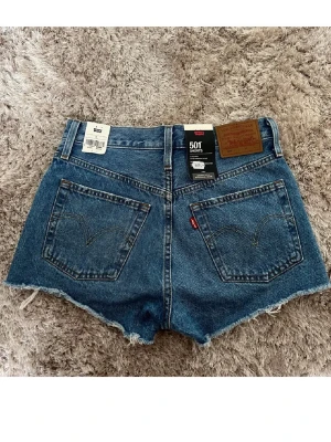 Levi's 501 blå jeansshorts - Säljer ett par klassiska Levi's 501 jeansshorts i blå denim med råa, fransiga benslut och slitna detaljer framtill. Shortsen har fem fickor, knappgylf och hög midja. Shortsen är helt oanvända med prislapp kvar!