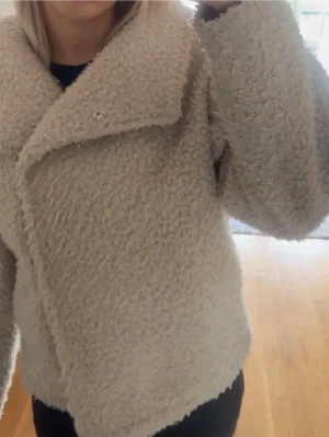 Teddy jacka - Supermjuk och fluffig teddyjacka från Gina trickot i beige färg. Jackan har bred krage, lång ärm och en oversized passform som ger en chill vibe. Perfekt för kalla dagar när du vill vara både varm och snygg.