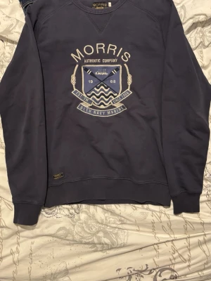 Marinblå sweatshirt från Morris - Säljer en marinblå sweatshirt från Morris med tryckt emblem och text på bröstet. Tröjan har rund halsringning, ribbade muddar och en relaxed passform. Perfekt för dig som gillar klassisk och stilren design.