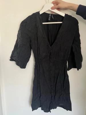 Svart kort omlottklänning zara  - Svart kort klänning från ZARA med v-ringning och omlottkänsla. Klänningen har korta ärmar, resår i midjan och är tillverkad i ett lätt tyg som ger en avslappnad look. Perfekt för dig som gillar enkel och stilren design.