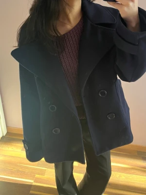 Mörkblå dubbelknäppt ullrock - Stilren mörkblå rock med dubbelknäppning och bred krage. Klassisk modell i ullmaterial som ger en snygg och tidlös look. Rocken har stora knappar och raka ärmar, perfekt för dig som vill ha en clean och trendig stil.