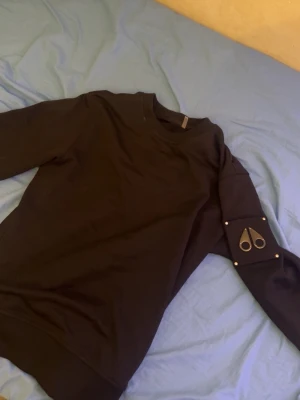 Svart hoodie från Moose Knuckles ÄKTA köpt på Nk Storlek S Lite stor i storleken - Säljer en svart hoodie från Moose Knuckles med deras ikoniska logga i metall på ena ärmen. Hoodien har rund halsringning, ribbade muddar och är gjord i mjukt bomullsmaterial. Perfekt för en clean och stilren look.