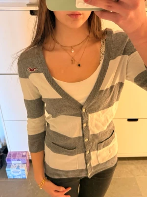 Vintage hollister randig gråvit  kofta med trekvarts ärmar  - Grå och vit kofta från Hollister med breda ränder och knappar framtill.                                                Storlek M men passar S bättre! Koftan har v-ringning, trekvarts ärmar och två fickor. Perfekt att slänga över en topp för en chill look.