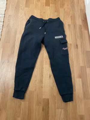 Svarta mjukisbyxor från Hoodrich - Säljer ett par svarta mjukisbyxor från Hoodrich med dragsko i midjan, fickor på sidorna och en extra ficka med lock på benet. Byxorna har tryckt logga på benet och är gjorda i mjukt, fleecefodrat material. Perfekta för chill dagar.