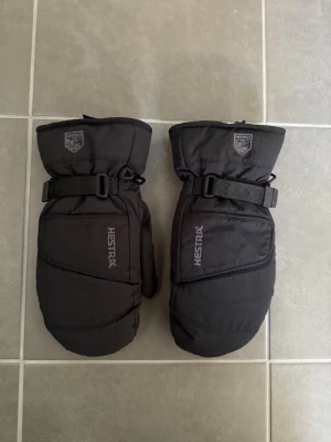 Svarta tumvantar från Hestra, stl 9 - Säljer ett par svarta tumvantar från Hestra med justerbara spännen runt handleden och logotyp på ovansidan. Vantarna är tillverkade i syntetmaterial och har en sportig design som passar perfekt för kalla vinterdagar.