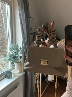 Beige handväska med scrunchie-handtag - Trendig beige handväska med scrunchie-inspirerat handtag och guldfärgad kedja. Väskan har ett lock med snyggt guldfärgat spänne och är tillverkad i slätt konstläder. Perfekt accessoar för att lyfta din outfit.